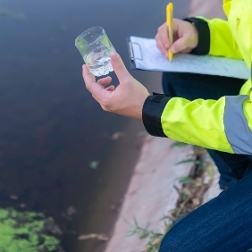 A researcher in a yellow reflective jacket taking notes while examining a test tube next to a canal.